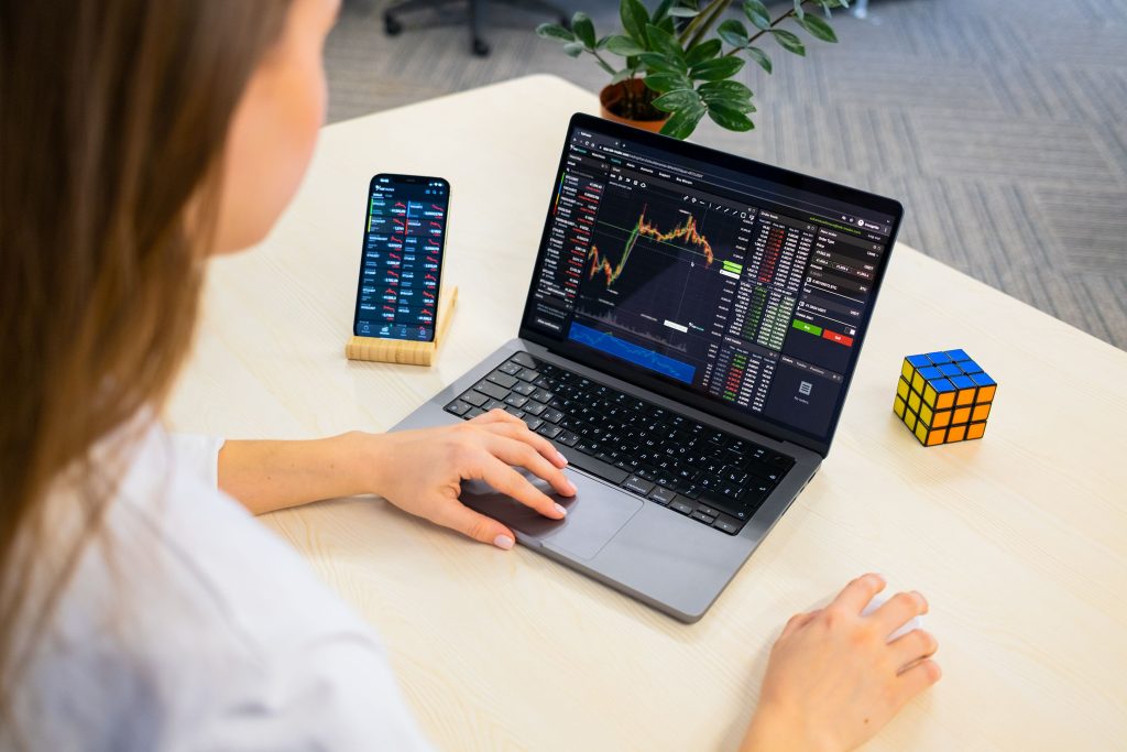 A woman using a laptop for stock trading analysis with a smartphone and Rubik's Cube nearby.